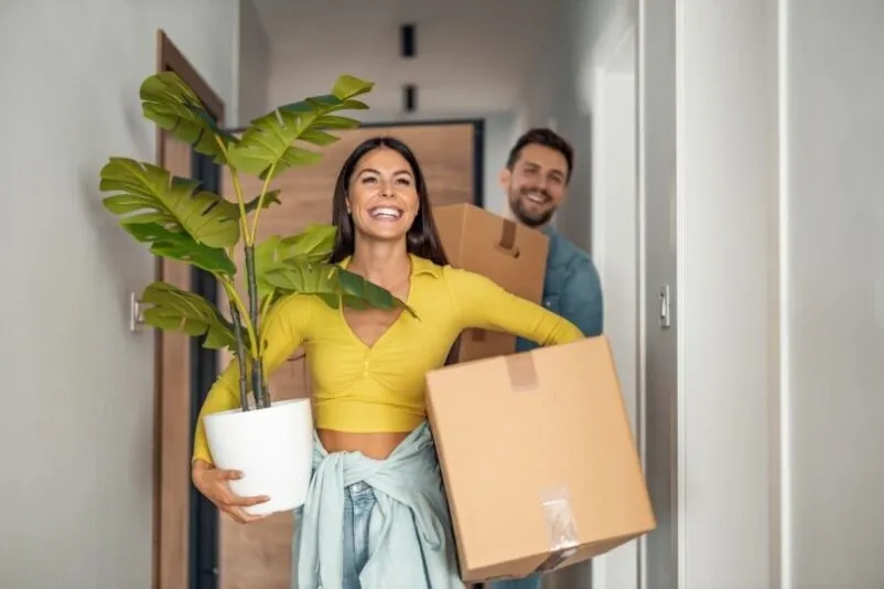 A young couple carry boxes and plants into their new home